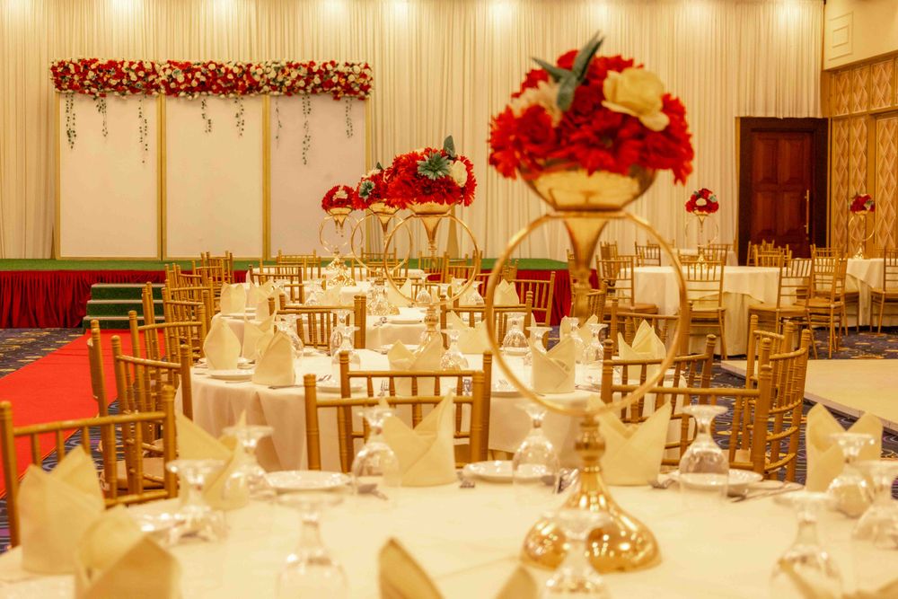 Round reception tables with tall red floral centrepieces and gold chairs before a draped stage
